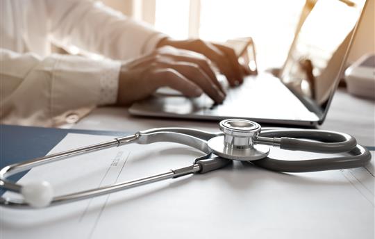 hospital records and stethoscope next to a doctor typing on a laptop 