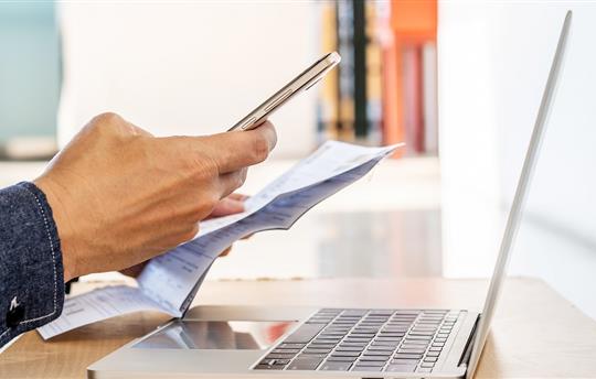A person holding a mobile phone in one hand and a paper statement in the other while sitting in front of an open  laptop