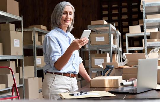 Female small business owner using mobile app checking parcel box and postal shipping order in warehouse